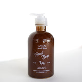 An 8 oz (473 mL) brown bottle of Ballad of the Bird Dog Hand Soap by Ballad of the Bird Dog, enriched with Santal essential oil and featuring a pump dispenser, sits on a white surface against a white background.