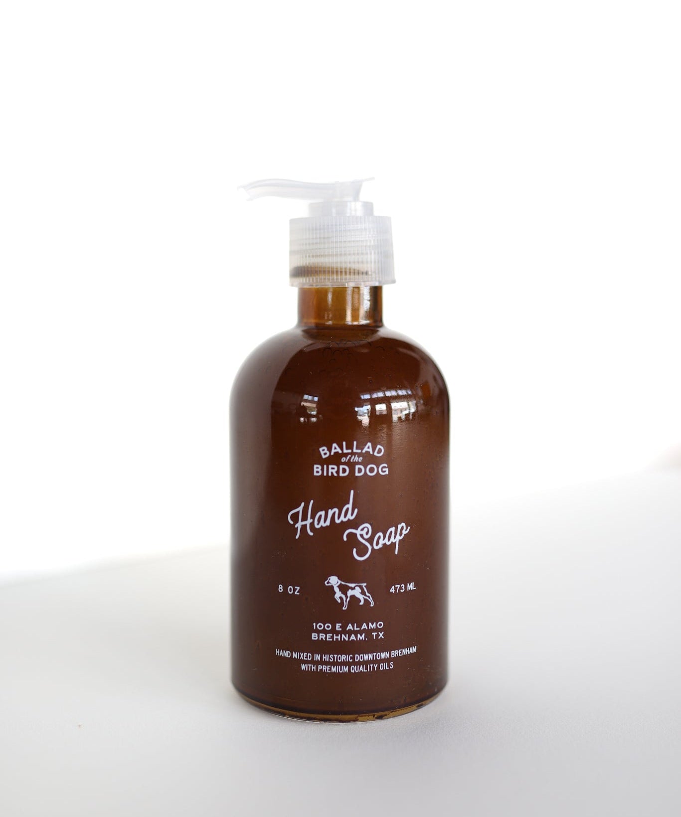 An 8 oz (473 mL) brown bottle of Ballad of the Bird Dog Hand Soap by Ballad of the Bird Dog, enriched with Santal essential oil and featuring a pump dispenser, sits on a white surface against a white background.