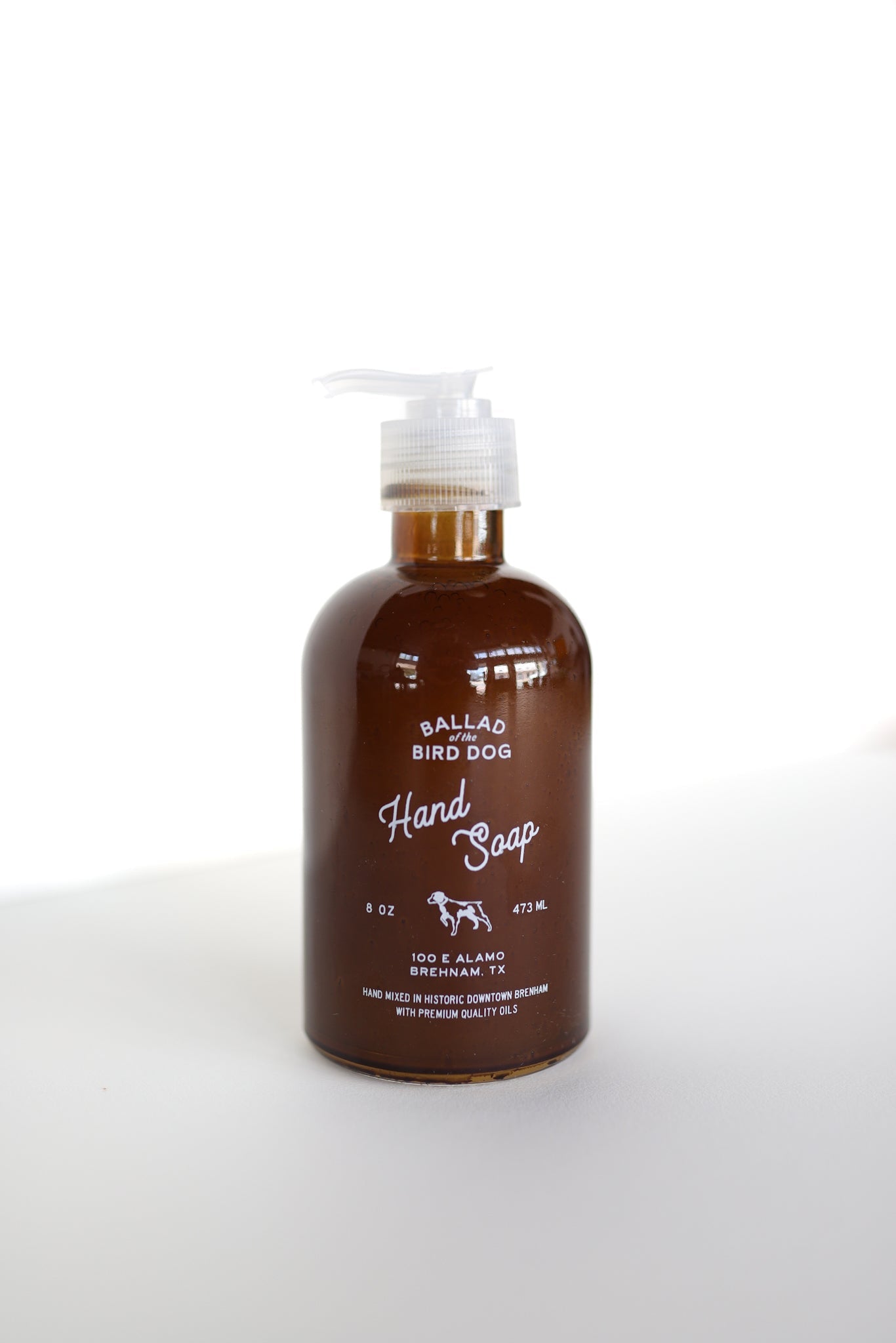 An 8 oz (473 mL) brown bottle of Ballad of the Bird Dog Hand Soap by Ballad of the Bird Dog, enriched with Santal essential oil and featuring a pump dispenser, sits on a white surface against a white background.