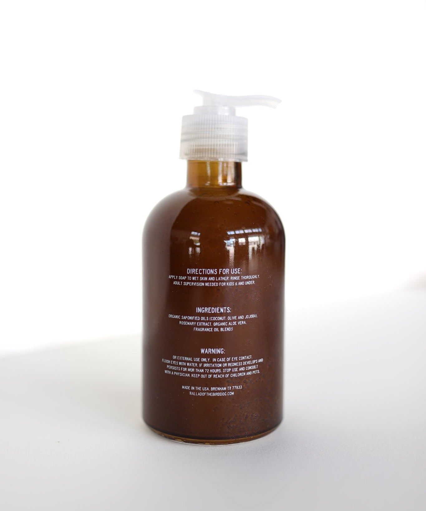 A brown pump bottle of Ballad of the Bird Dog Hand Soap with white text showing directions, Santal essential oil ingredients, and warnings stands on a white surface against a bright background.