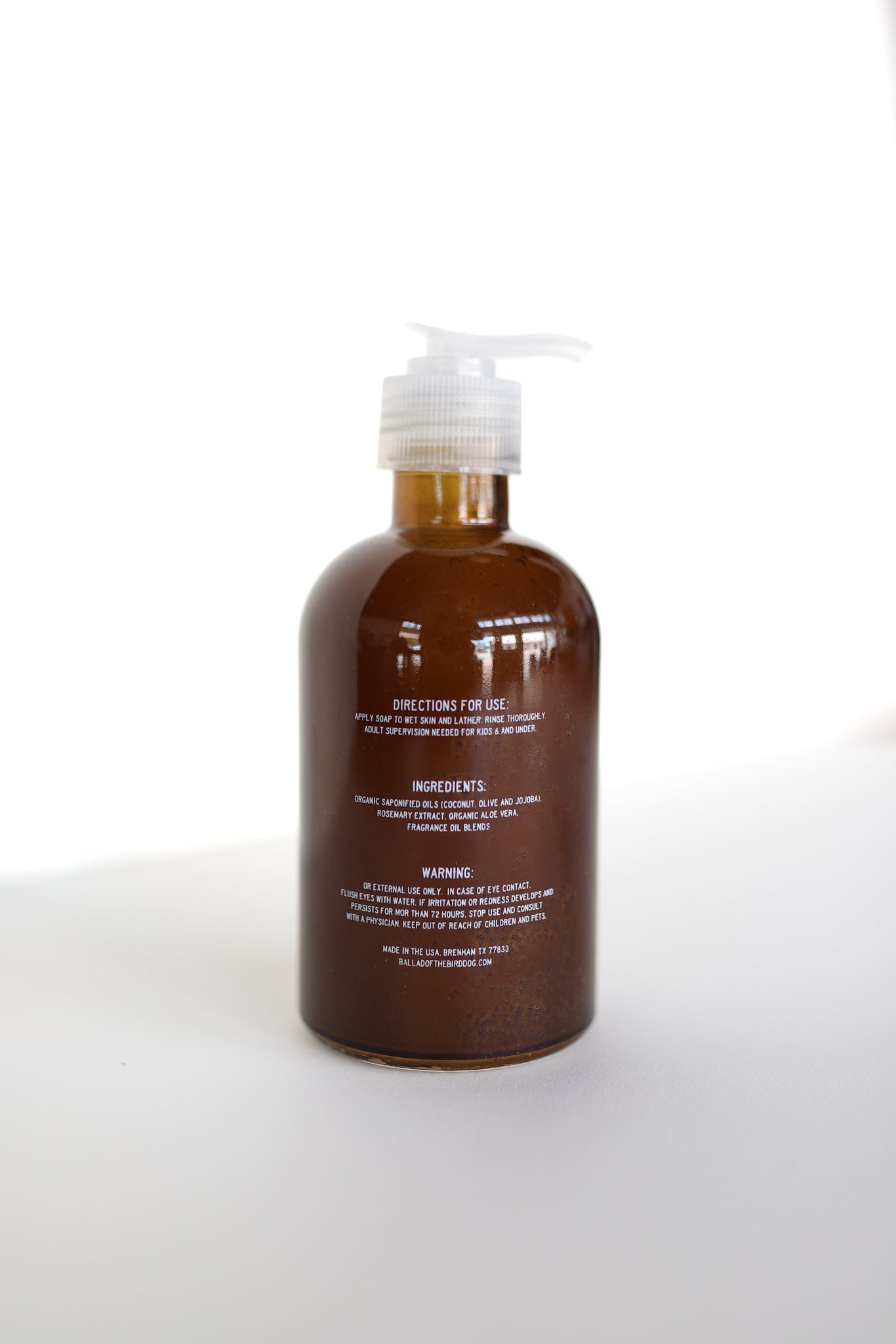 A brown pump bottle of Ballad of the Bird Dog Hand Soap with white text showing directions, Santal essential oil ingredients, and warnings stands on a white surface against a bright background.