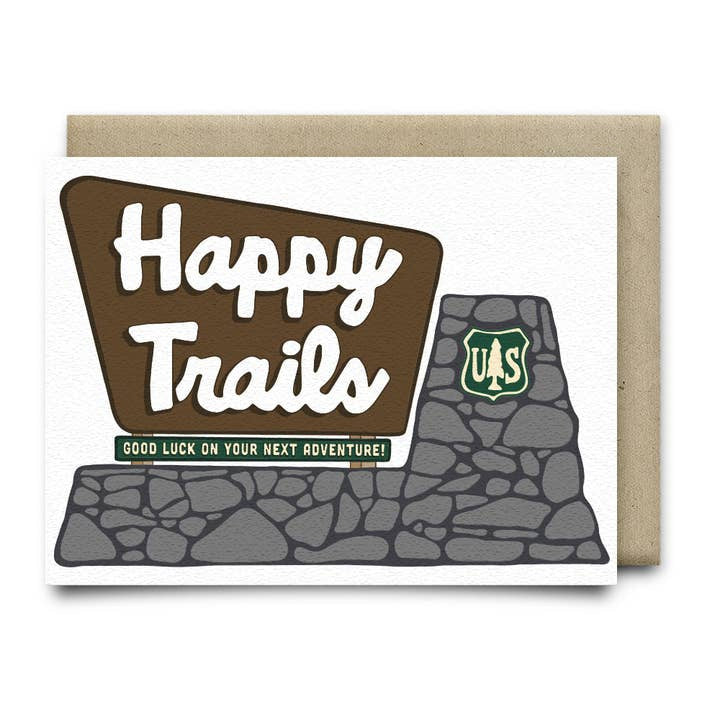 The Happy Trails - Good Luck Card by Anvil Cards features a hand-drawn stone sign with "Happy Trails" and "Good luck on your next adventure!" on sustainable linen paper, plus a green U.S. Forest Service emblem.