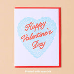 The Happy Valentines Day Card by And Here We Are features a blue lace heart, vintage “Happy Valentine’s Day” text, neon ink, a red border, and light beige background.