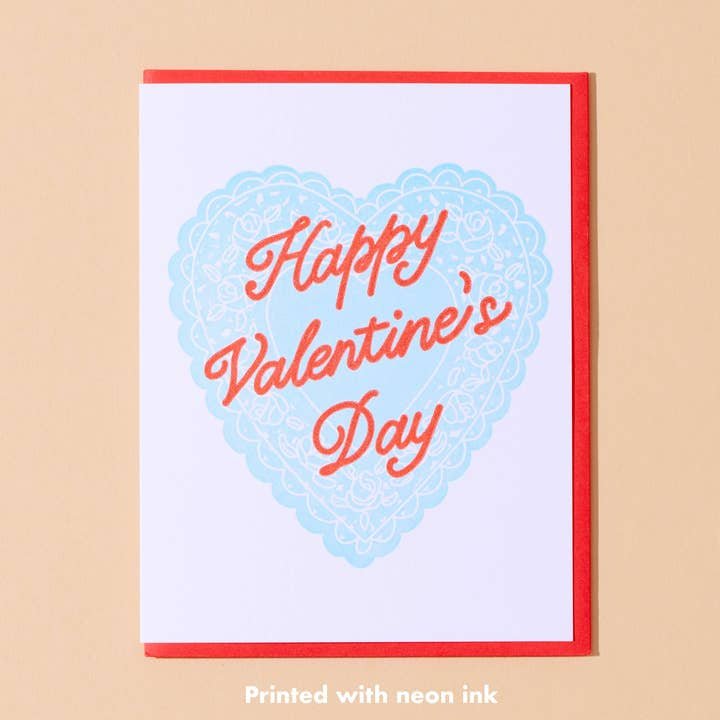 The Happy Valentines Day Card by And Here We Are features a blue lace heart, vintage “Happy Valentine’s Day” text, neon ink, a red border, and light beige background.