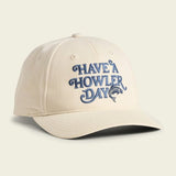 The Have a Howler Day cap by Howler Brothers features beige fabric, blue "HAVE A HOWLER DAY" embroidery above a jumping fish, classic Howler artwork, and a comfy snapback closure for a perfect fit.