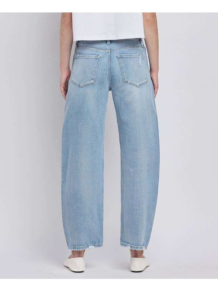 Person standing with back to camera, wearing Vervet High Rise Light Wash Barrel Leg Jeans, a white top, and white flat shoes against a plain background.