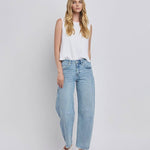 A woman stands against a plain background wearing a sleeveless white top, Vervet High Rise Light Wash Barrel Leg Jeans with a relaxed fit, and white flat shoes.