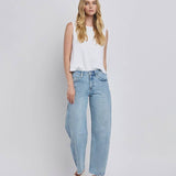 A woman stands against a plain background wearing a sleeveless white top, Vervet High Rise Light Wash Barrel Leg Jeans with a relaxed fit, and white flat shoes.