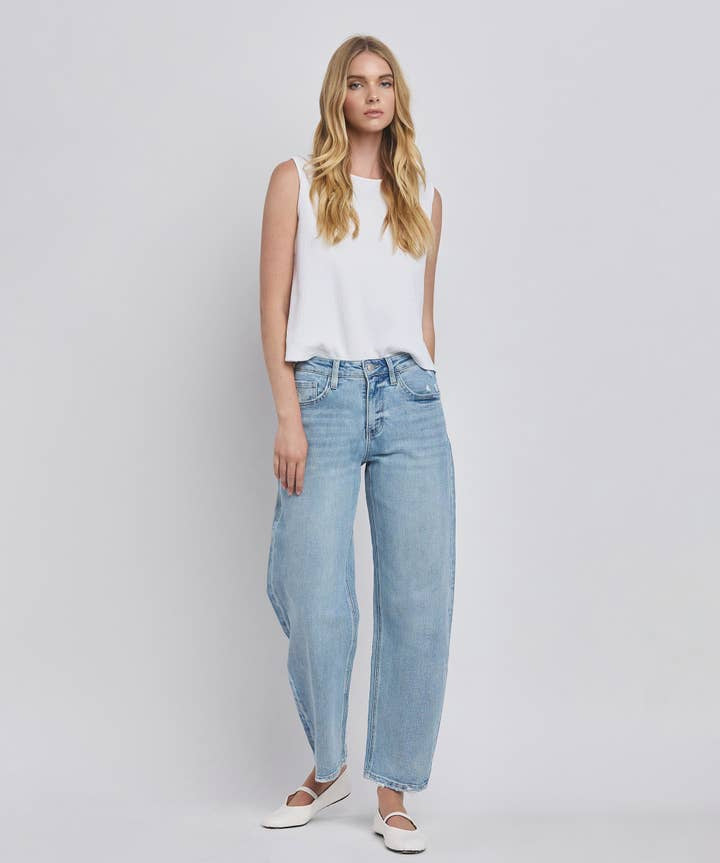 A woman stands against a plain background wearing a white sleeveless top, Vervet High Rise Light Wash Barrel Leg Jeans, and white flats.