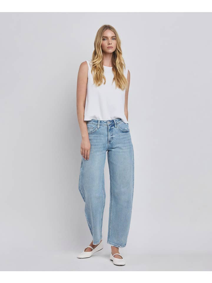 A woman stands against a plain background wearing a white sleeveless top, Vervet High Rise Light Wash Barrel Leg Jeans, and white flats.