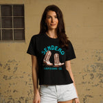 A woman stands indoors against a beige wall, wearing the Sendero Provisions Co. High Stepper Boxy Tee with a cowboy boot graphic and white denim shorts.