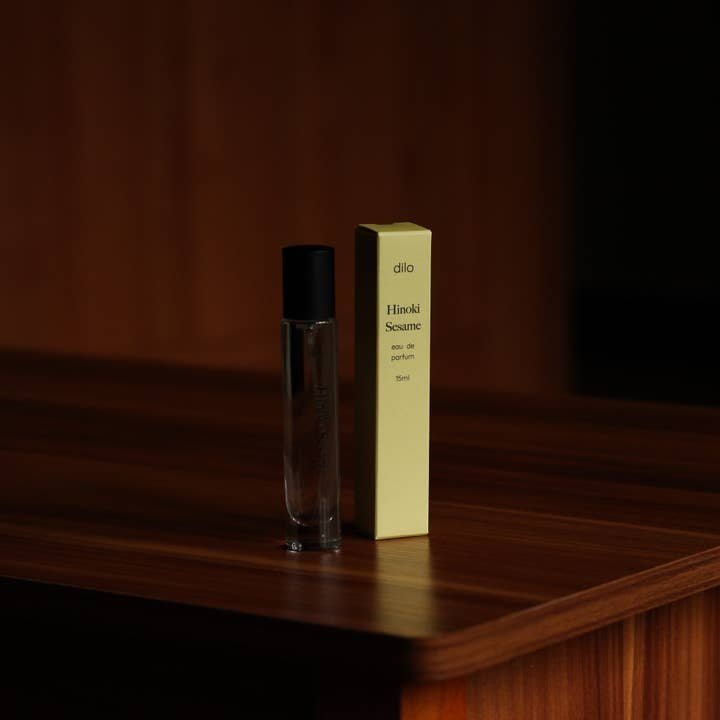 A clear glass bottle of Hinoki Sesame Eau De Parfum by dilo with a black cap sits beside its yellow box on a wooden surface in dim lighting, evoking sandalwood and sesame notes.