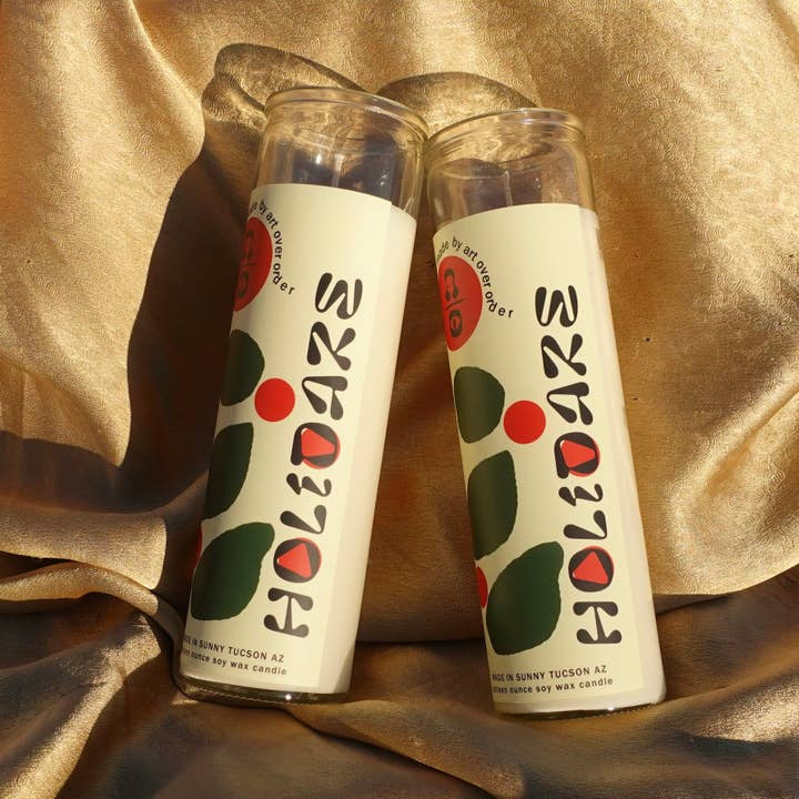 Two Holidaze Intention Candles by Art Over Order in glass jars rest on gold satin, with warm green cardamom notes infusing the scene with a spicy fragrance.