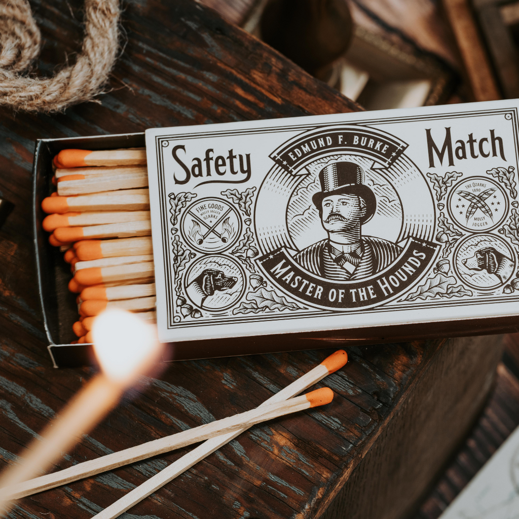 An open box of Mollyjogger Hounds Safety Matches with orange tips sits on a wooden surface; one match is lit in the foreground.