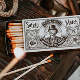 An open box of Mollyjogger Hounds Safety Matches with orange tips sits on a wooden surface; one match is lit in the foreground.