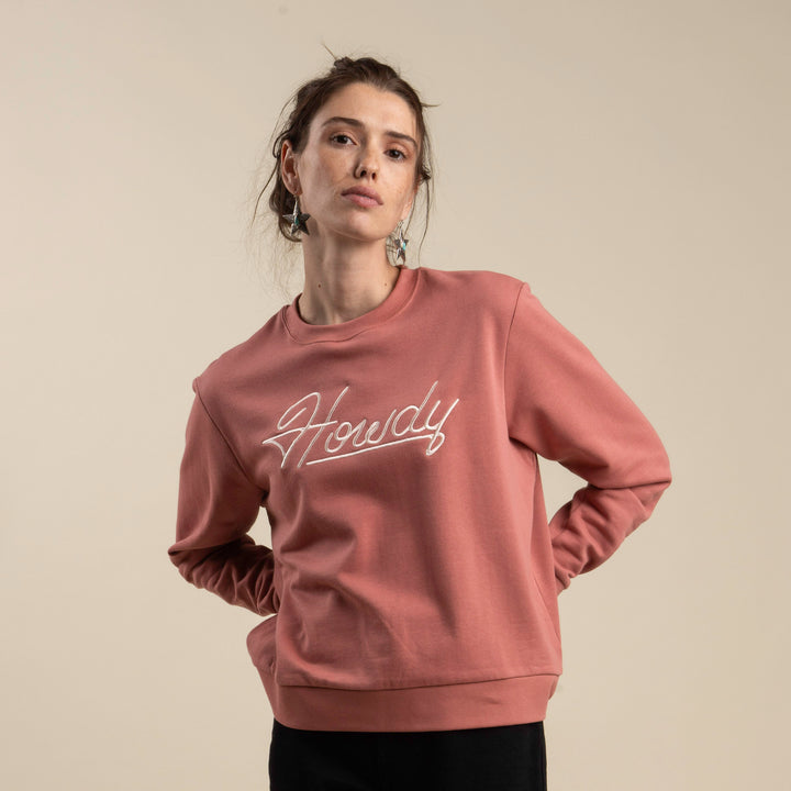 A person faces the camera wearing the Sendero Provisions Co. Howdy Drop Shoulder Sweater, an ultra-soft dusty pink sweatshirt with “Howdy” in cursive on the front.
