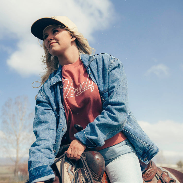A person in a Sendero Provisions Co. Howdy Drop Shoulder Sweater, denim jacket, and cap rides a horse outdoors under a blue sky.