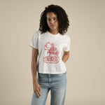 A person in Sendero Provisions Co.’s Howdy Stranger Boxy Tee—a soft, white organic cotton shirt with a red "Howdy Stranger" graphic—and light blue jeans stands against a beige background.