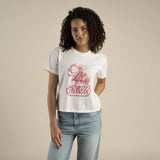 A person in Sendero Provisions Co.’s Howdy Stranger Boxy Tee—a soft, white organic cotton shirt with a red "Howdy Stranger" graphic—and light blue jeans stands against a beige background.