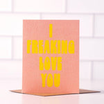 The I Freaking Love You card by Daydream Prints features bold yellow text on a pink background, shown standing upright on a white surface with a blurred tiled background.