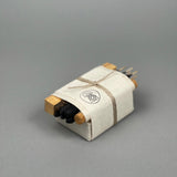 The Bath & Meditate Bundle by Incausa is a small set wrapped in white Nepalese Lokta paper and tied with string, containing charcoal, cork, and wooden sticks, displayed on a gray background.