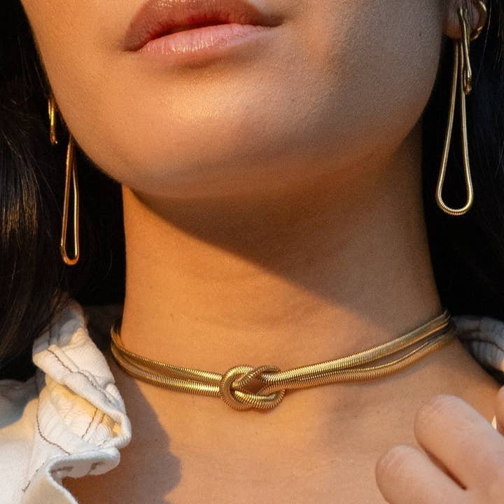 A woman wears the Peter and June In The Loop Choker, featuring a knot design, paired with 18k gold plated teardrop earrings and a white garment.