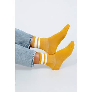 Model wears the Jouer Ankle Sock by Tailored Union in yellow with white stripes, styled with light blue jeans and feet extended against a plain light background.