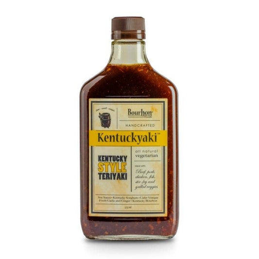 A clear glass bottle of Kentuckyaki by Bourbon Barrel Foods, a sorghum-sweetened Kentucky Bourbon teriyaki sauce, with a gold cap and beige label.