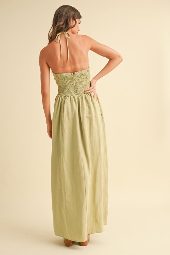 A woman stands facing away, wearing the Knotted Front Halter Neck Dress by Miou Muse—a light green, sleeveless linen-cotton backless maxi with smocked bodice in size S, ideal for elegant women's clothing collections.