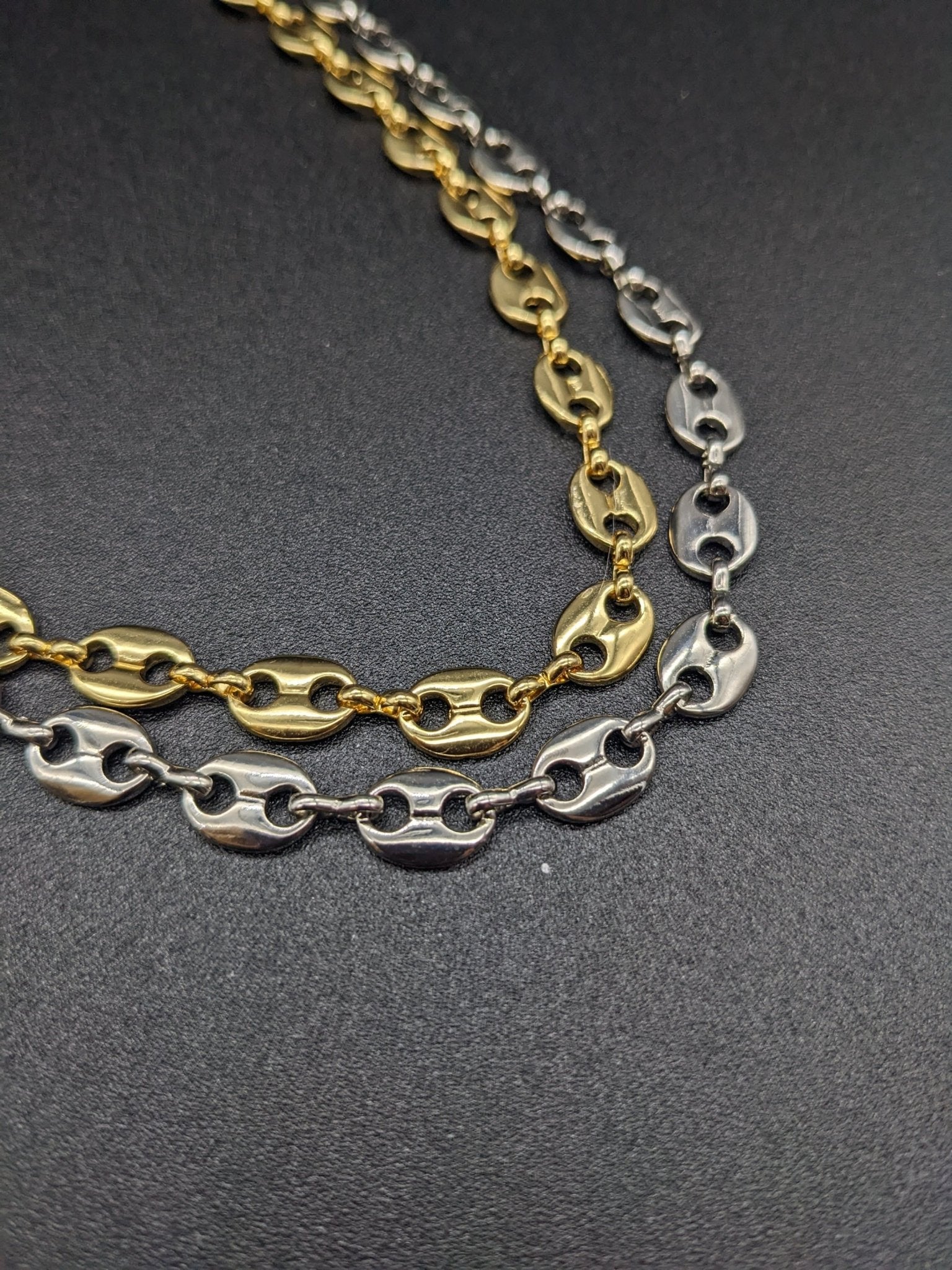 The Koffee Necklace by Minh Atelier, featuring two chain necklaces with oval links—one gold plated choker and one silver plated necklace—is displayed on a plain black surface.