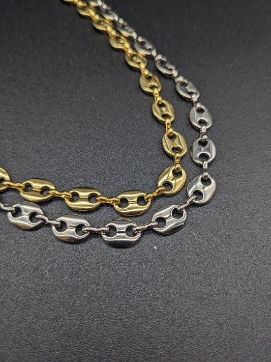 The Koffee Necklace by Minh Atelier, featuring two chain necklaces with oval links—one gold plated choker and one silver plated necklace—is displayed on a plain black surface.