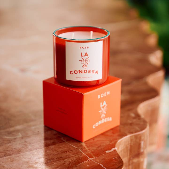 A red glass La Condesa | Roen candle by Roen, made from coconut soy wax, rests on a matching branded box on a polished stone surface.