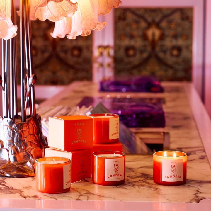 Three lit La Condesa | Roen candles by Roen, made with coconut soy wax, are shown in matching boxes on a marble counter next to a lamp with pink lampshades.