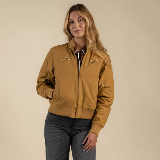 A woman stands against a beige background, one hand in her pocket, wearing Sendero Provisions Co.'s La Tierra Jacket—a tan cotton canvas piece with embroidered details.