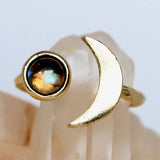The Mesa Blue Labradorite Crescent Ring features a gold-plated adjustable band with a round iridescent labradorite stone on one end and a crescent moon shape on the other, shown on a light mineral background.