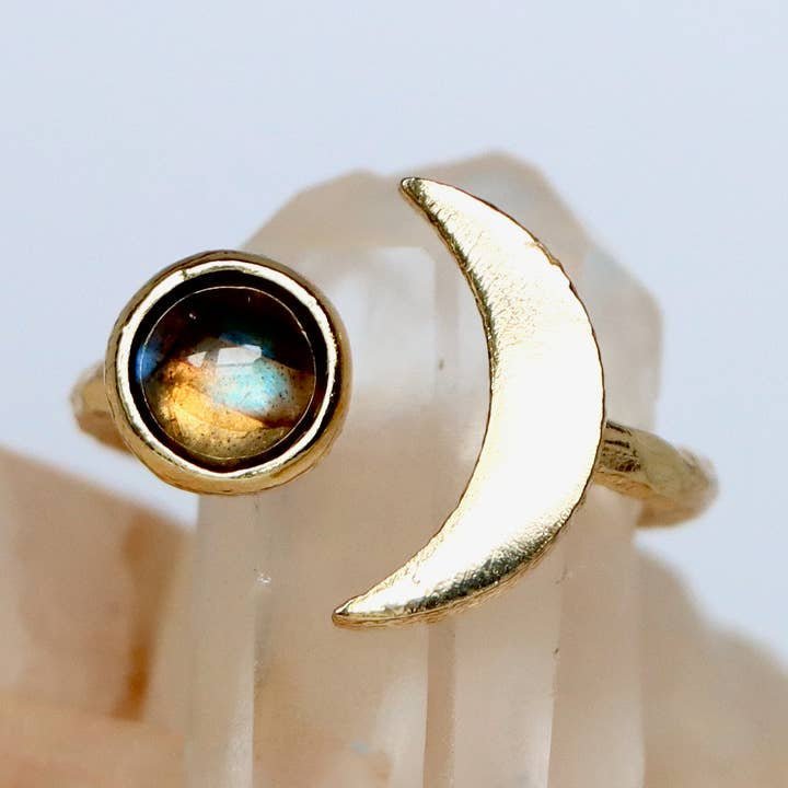 The Mesa Blue Labradorite Crescent Ring features a gold-plated adjustable band with a round iridescent labradorite stone on one end and a crescent moon shape on the other, shown on a light mineral background.