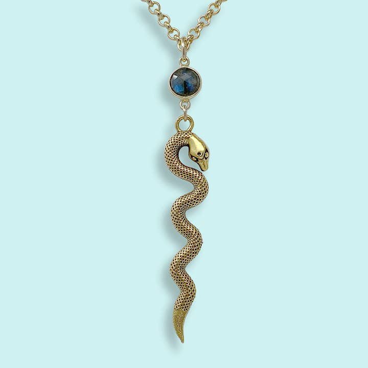 The Labradorite Serpent Necklace by Ornamental Things features a textured gold snake charm and a round labradorite pendant on a gold rolo chain, shown against a light blue background.