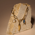 The Peter and June Lagoon Charm Bracelet is a waterproof 18k gold-plated chain adorned with pearl, seashell, fish, and aquamarine gemstone charms, displayed on a textured beige stone.