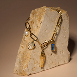 The Peter and June Lagoon Charm Bracelet is a waterproof 18k gold-plated chain adorned with pearl, seashell, fish, and aquamarine gemstone charms, displayed on a textured beige stone.
