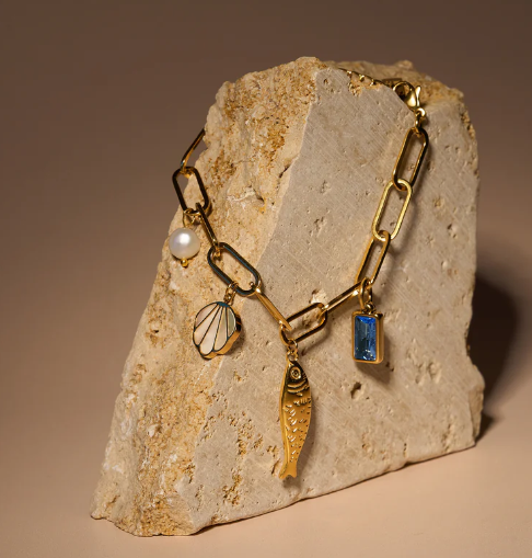 The Peter and June Lagoon Charm Bracelet is a waterproof 18k gold-plated chain adorned with pearl, seashell, fish, and aquamarine gemstone charms, displayed on a textured beige stone.