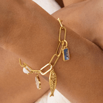 The Lagoon Charm Bracelet by Peter and June is an 18k gold plated chain with large links, adorned with aquamarine charms, a fish, and other small ornaments.
