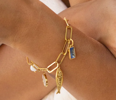 The Lagoon Charm Bracelet by Peter and June is an 18k gold plated chain with large links, adorned with aquamarine charms, a fish, and other small ornaments.