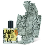 A square glass bottle labeled "Lampblack | Fzotic" stands before its black-and-white patterned, unfolded box, reflecting the fragrance’s distinctive woody-smoky character.
