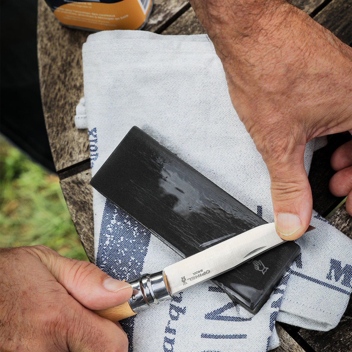 A person sharpens a knife with the Opinel Large Natural Sharpening Stone, using a cloth on a wooden surface for optimal knife maintenance.