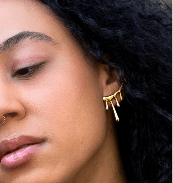 Close-up of a person with curly dark hair wearing the Lava Studs by Peter and June, 18k gold plated earrings with a dripping design, along with a small nose ring.
