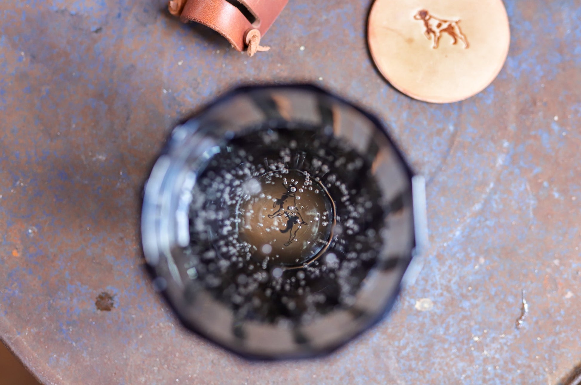 Top-down view of a glass with fizzy, dark liquid next to the Ballad of the Bird Dog Leather Coaster 4 Piece Set—handmade coasters embossed with a dog, perfect for stylish desk protection.