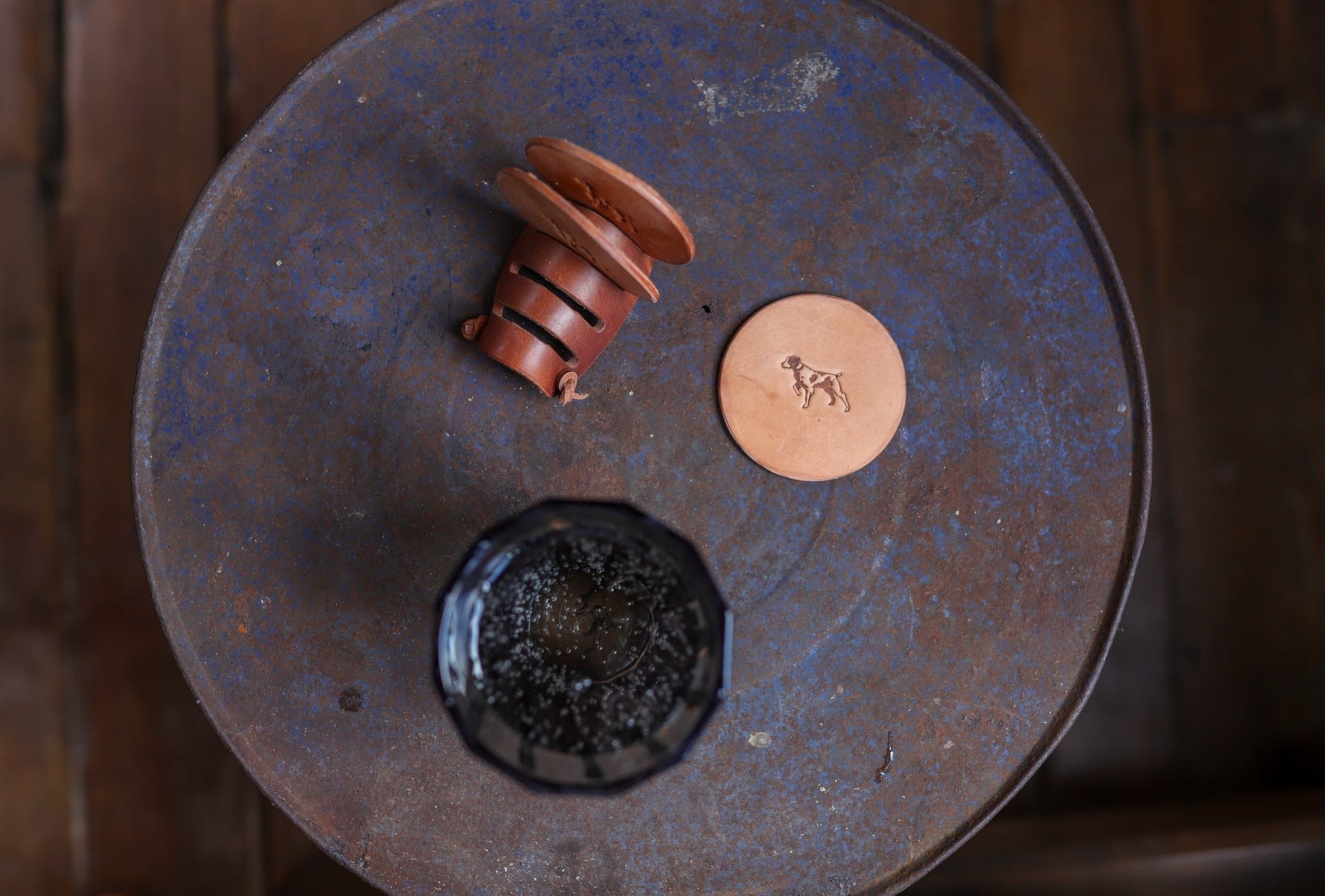 A round metal table with dark glass, a brown ceramic cup on its side, and Ballad of the Bird Dog’s Leather Coaster 4 Piece Set—handmade round leather coasters with dog engraving for stylish desk protection.