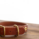 The Leather Dog Collar by Ballad of the Bird Dog, crafted from rich brown leather with brass hardware, rests on wood and highlights the brand's quality craftsmanship.