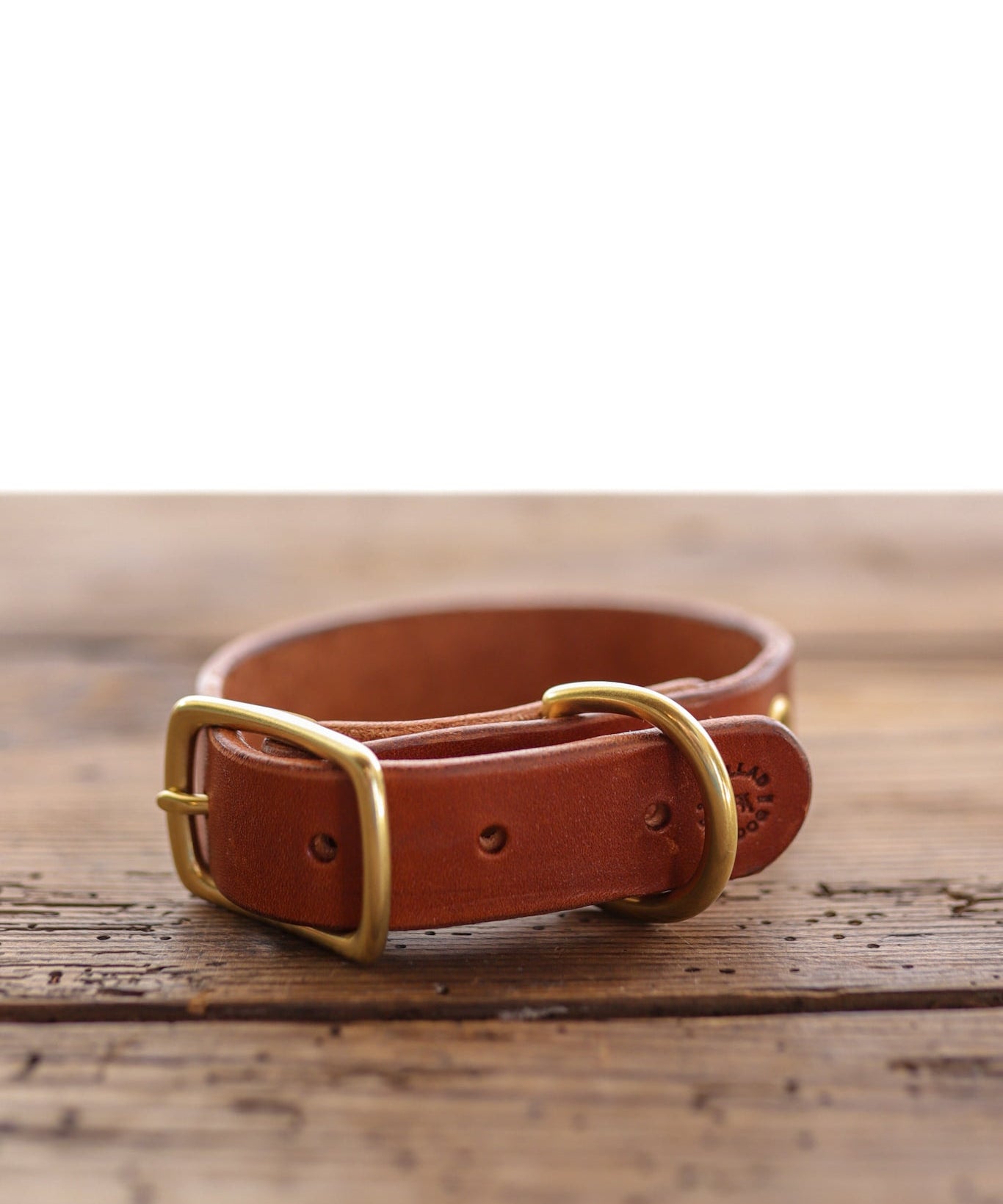 The Leather Dog Collar by Ballad of the Bird Dog, crafted from brown English bridle leather with a gold-colored buckle and D-ring, is displayed on a wooden surface.