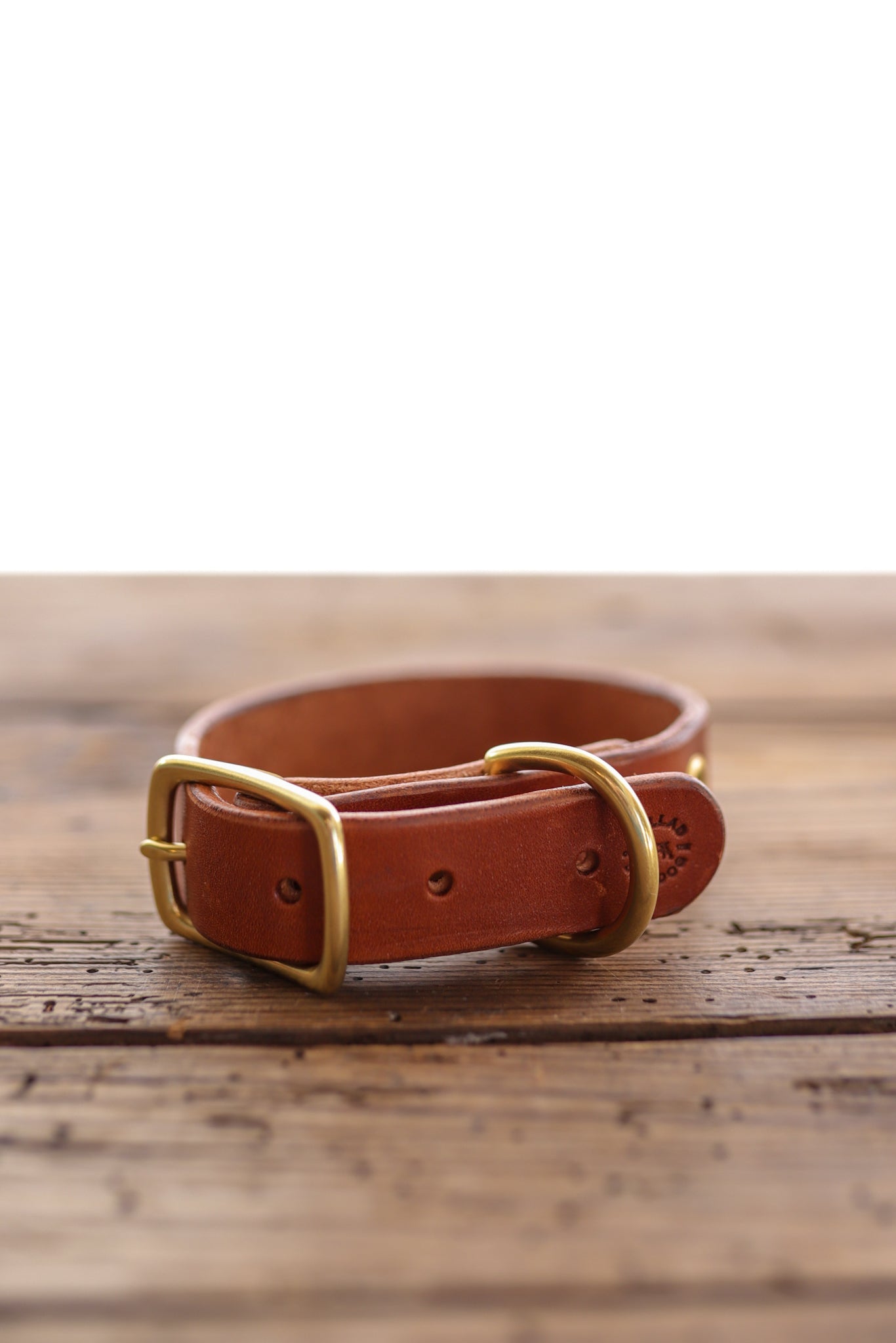 The Leather Dog Collar by Ballad of the Bird Dog, crafted from brown English bridle leather with a gold-colored buckle and D-ring, is displayed on a wooden surface.
